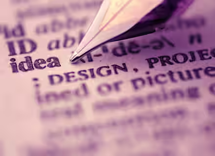 A close-up image of a fountain pen tip pointing at the word "idea" in a dictionary. The surrounding text is slightly blurred, emphasizing the word "idea". The scene is bathed in a subtle, warm, purple hue.