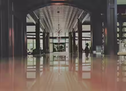 A modern, upscale bar with a long, reflective counter leading to a large window at the back. Wooden paneling and contemporary chandeliers adorn the ceiling. Silhouettes of people are visible, creating a sophisticated, atmospheric setting.