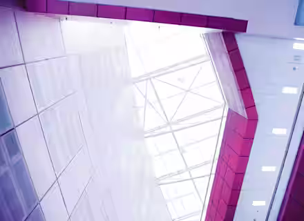A view looking up at the geometric ceiling of a modern building, featuring large, angled glass panels that allow natural light to flood the interior. The surrounding walls are clad in red and white tiles, creating a colorful, contemporary architectural design.