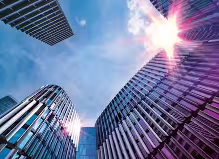 A low-angle view of modern office buildings with reflective glass and metal facades, rising towards a bright sky. The sun is shining brightly, creating lens flare effects on the buildings. The architectural structures are sleek and contemporary.
