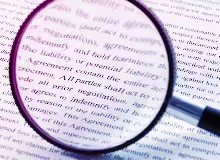 A magnifying glass is focused on a printed document, enlarging and highlighting specific legal terms and phrases, such as "liability" and "All parties shall act," indicating close scrutiny of the text. The background is out of focus, showing more text.