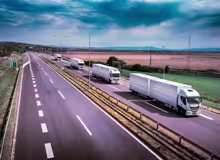 A fleet of white semi-trucks drives on a multi-lane highway through a rural area. The sky is overcast, and fields stretch out on either side of the road. Street lights line the highway, and distant mountains are visible on the horizon.