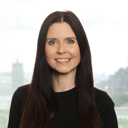 A woman with long, straight dark brown hair is smiling at the camera. She is wearing a black short-sleeve shirt and stands in front of a window with a blurry cityscape background.