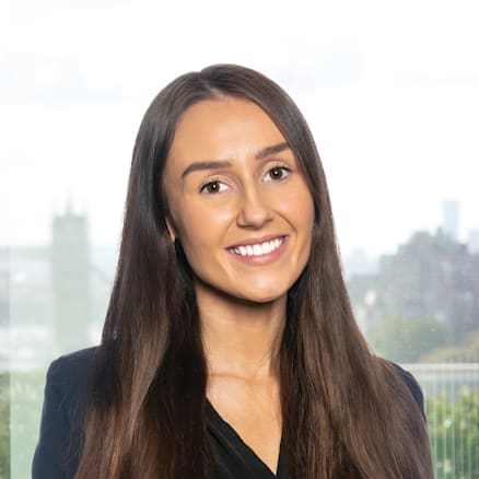 A woman with long brown hair wearing a black blazer and black shirt is smiling in front of a window with a blurry cityscape and greenery outside.