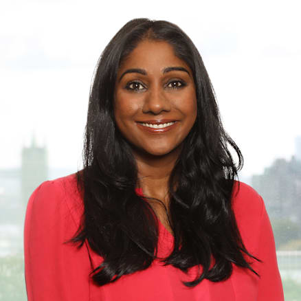 A woman with long black hair, wearing a bright red blouse, smiles while standing in front of a blurred outdoor background.