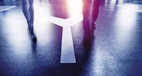 Two people are walking on a wet street at night, illuminated by streetlights. They are approaching a large painted Y-shaped arrow on the ground, which splits into two directions. The scene has a moody, reflective atmosphere.
