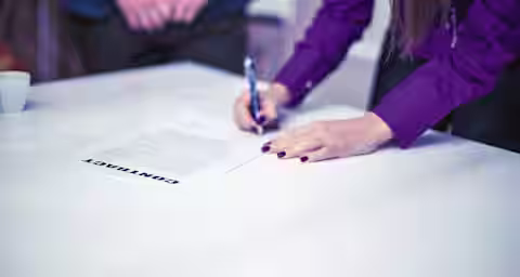 A person wearing a purple shirt is signing a contract on a white table with a pen. Another person in a blue suit stands opposite, with only their torso and hands visible. A small white coffee cup is placed on the table in the background.