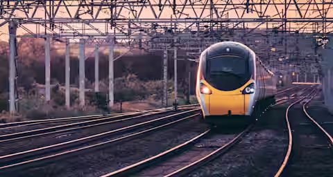A modern yellow and black train travels along a set of railway tracks at dusk. The train is emerging from beneath a network of overhead electrical wires, surrounded by trees and rural landscape in the background.