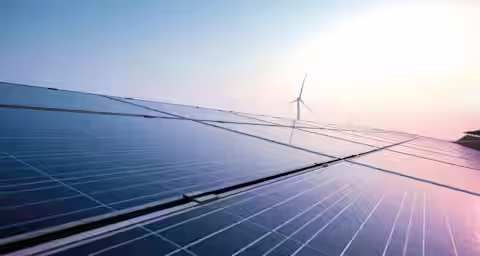 Close-up of solar panels with a wind turbine in the background under a clear sky. The sun is shining brightly, reflecting off the panels, representing renewable energy sources.