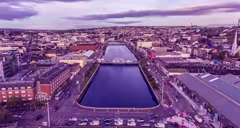 Aerial view of a city with a river running through the center, surrounded by buildings and roads, with a bridge crossing the river and cars parked along the streets under a cloudy, purple-tinged sky.