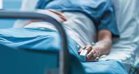 A person lies in a hospital bed wearing a blue gown, with one hand resting beside them holding a medical finger clip, possibly a pulse oximeter. The focus is on the lower part of their body, highlighting the medical equipment and hospital surroundings.