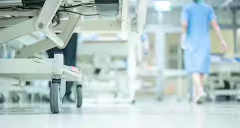 Close-up of a hospital bed's wheels on a polished floor. A medical professional in blue scrubs is seen walking away in the background, with blurred hospital equipment and lights creating a clinical atmosphere.