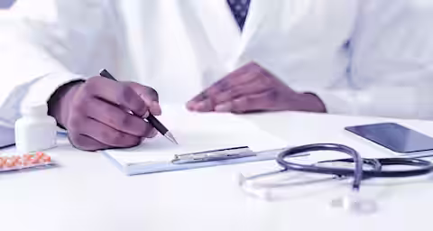 A doctor wearing a white coat and a tie writes on a clipboard. In the foreground, there are a stethoscope, a smartphone, and medication bottles with pills blister packs on the desk. The background is blurred.
