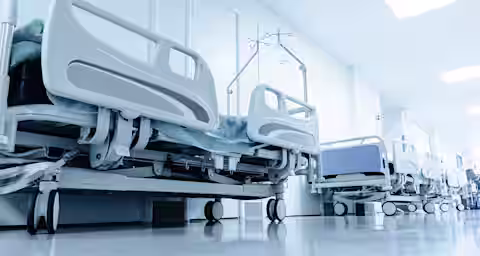 A row of empty hospital beds with blue sheets lined up against a white wall in a brightly lit, sterile hospital corridor. The floor is shiny, and there are IV stands next to each bed.