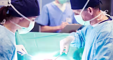 Surgeons in blue scrubs and face masks are performing an operation in a sterile operating room. A nurse in the background takes notes while observing the procedure. Bright surgical lights illuminate the scene.