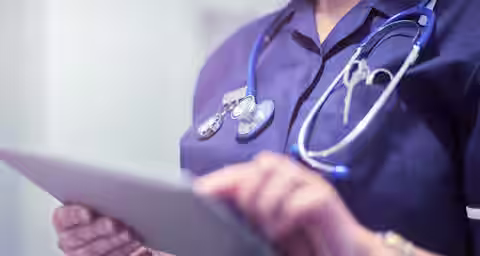 A healthcare professional wearing a dark blue uniform with a stethoscope around their neck is holding and using a tablet. The image focuses on their chest and hands, obscuring their face. The background is blurred, highlighting the individual.