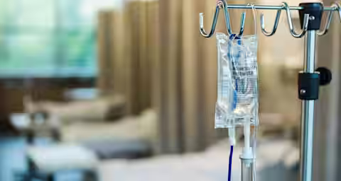 An IV drip bag hangs from a stand in a hospital room. The background shows several empty hospital beds with beige curtains separating them. The scene is softly lit, suggesting a calm, clinical environment.