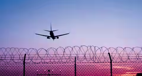 A commercial airplane is seen landing at an airport during sunset. The silhouette of the plane is prominent against a colorful sky of pink, purple, and blue hues. In the foreground, a barbed wire fence and security lights are visible.