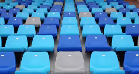An empty stadium seating area with rows of plastic seats. The seats are predominantly blue, with a few gray ones interspersed randomly. Small white tags are visible on the backs of each seat.