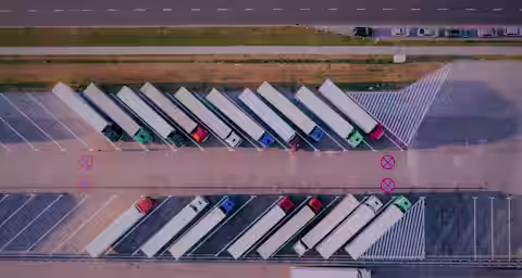 Aerial view of a parking lot with several semi-trucks parked diagonally in designated spaces. A single truck is parked perpendicularly at the lot's edge. There are red "X" markings indicating no-parking zones, and a road with moving vehicles is visible nearby.