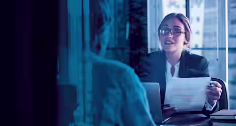 A woman wearing glasses and a business suit is holding a paper and speaking to another person across a desk in an office setting. The scene is viewed through a window, reflecting the cityscape outside.