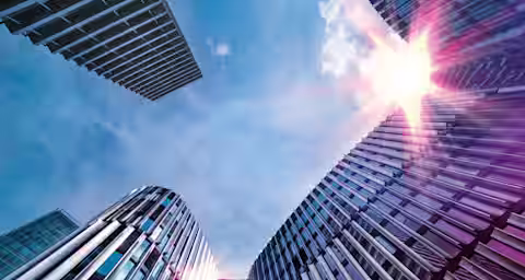 A low-angle view of modern office buildings with reflective glass and metal facades, rising towards a bright sky. The sun is shining brightly, creating lens flare effects on the buildings. The architectural structures are sleek and contemporary.
