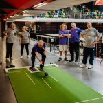 Image shows multiple people playing short mat bowls with a leisure centre sports hall in the background