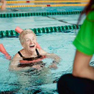 picture shows an older lady using a swimming aid looking happy at the edge of a pool with a lifeguard watching