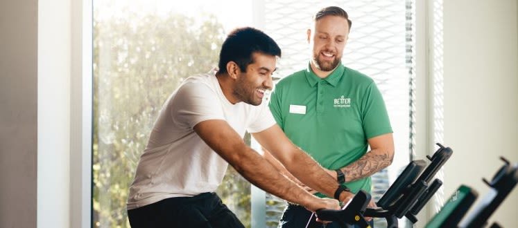 image shows a male on an exercise bike with an instructor supporting