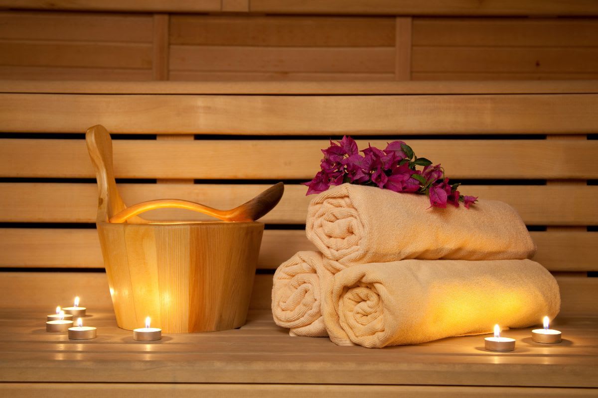Saunazubehör bestehend aus einem Holzkübel, einer Kelle, aufgerollten Handtüchern und Teelichtern auf einer Holzbank in der Sauna.