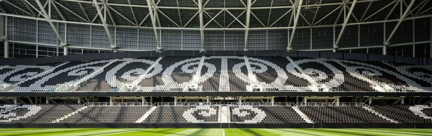 Empty stadium with a green grass field and black and white patterned seating under a white steel roof structure.