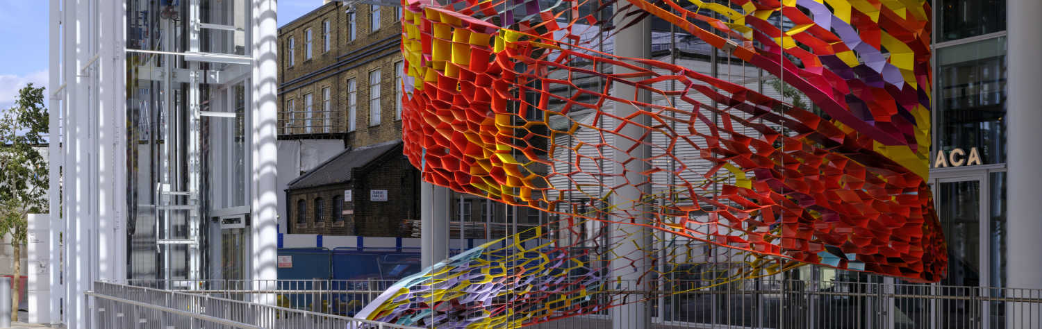A large, colorful, abstract sculpture made of interconnected geometric shapes hangs from a modern building's ceiling.