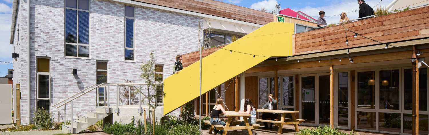 Modern two-story building with white brick walls, a yellow architectural feature, and people on a rooftop deck.