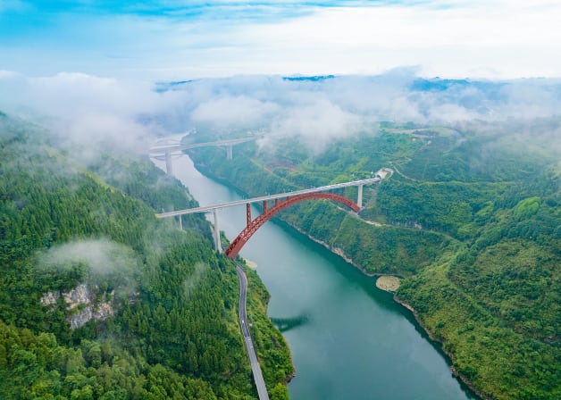 A river with a bridge with a red arch connecting the mountains across it.