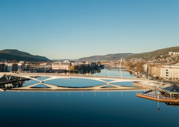 A modern white pedestrian bridge spans a calm blue river, reflecting the bridge and distant buildings under a clear sky.