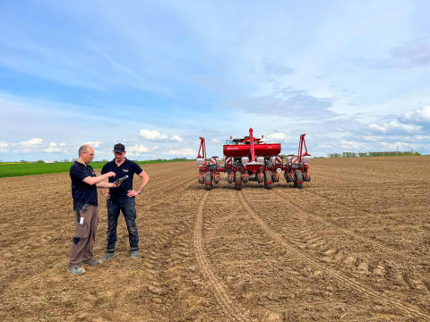 Two men discussing in a field during Kverneland Optima TFprofi PUDAMA trials, with the red precision seeder positioned behind them on freshly cultivated soil, showcasing advanced seed and fertilizer placement technology.