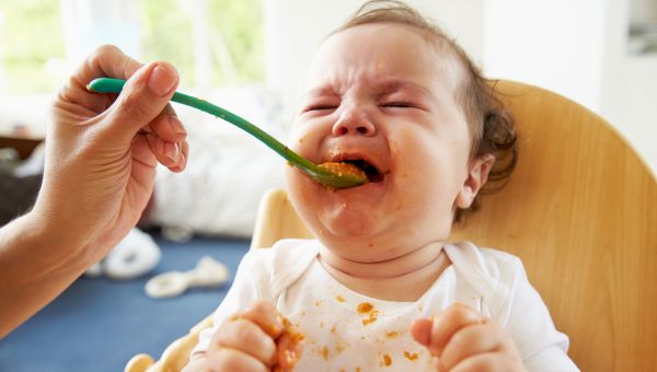 Mother feeding infant spoonfuls of baby food while baby cries