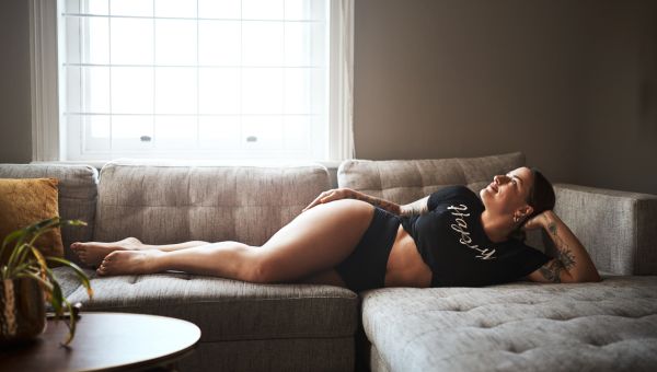 a body-positive young White woman with tattoos lies on a couch in her underwear with a smile