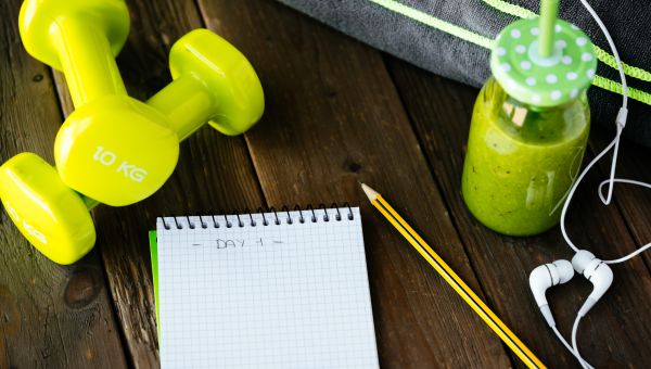a closeup shot of a table with a workout log notebook, dumbbells, and a green smoothie