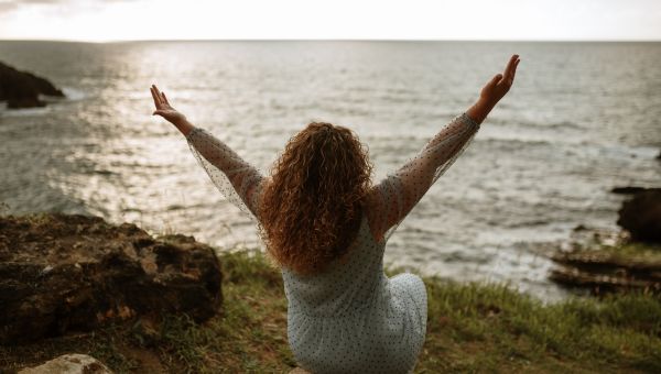 Back view of a pregnant person holding both hands up while looking out at the ocean