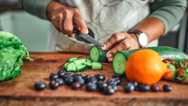 View of an unidentifiable man cutting up cucumber on a cutting board with other vegetables and fruits on the counter