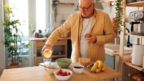 an older man with white hair and glasses pours fresh orange juice into a glass in his kitchen with a bowl of strawberries, blueberries and bananas on the counter