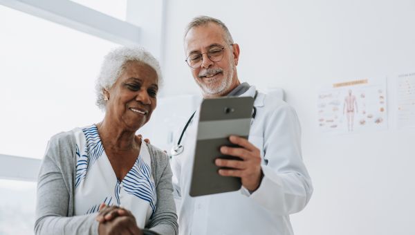 an older Black woman consults with her healthcare provider, an older white man; he shows her an image on a digital tablet