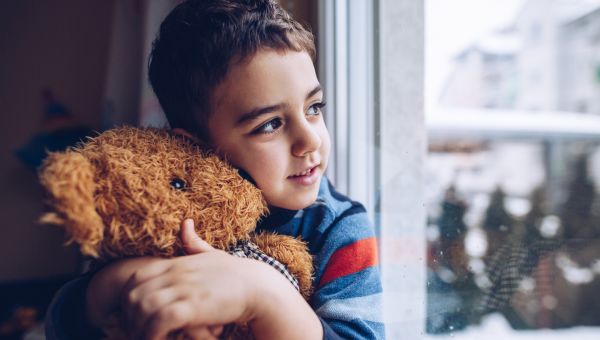 A young boy hugging his teddy bear near a window