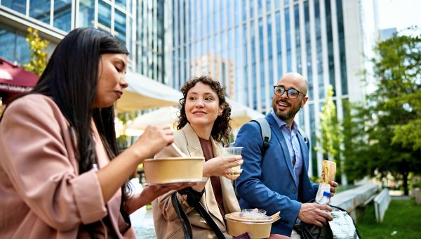three office workers, a Latina woman, a white woman, and a Black man, enjoy eating lunch outside on a spring day