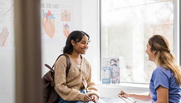 a young Black woman smiles as she receives preventive health care from a woman nurse practitioner