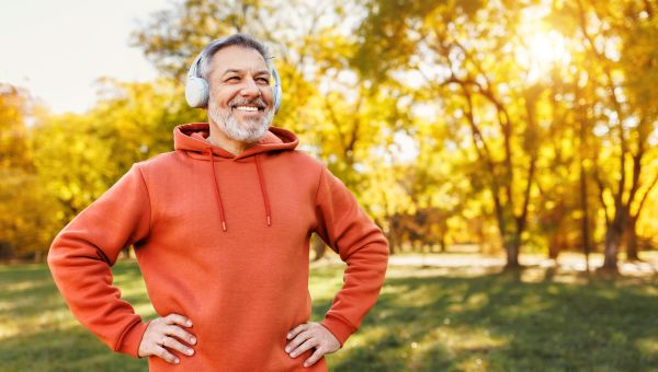 a smiling middle aged Hispanic man with a beard wears earphones and a red sweatshirt as he gets ready to exercise outdoors in a park