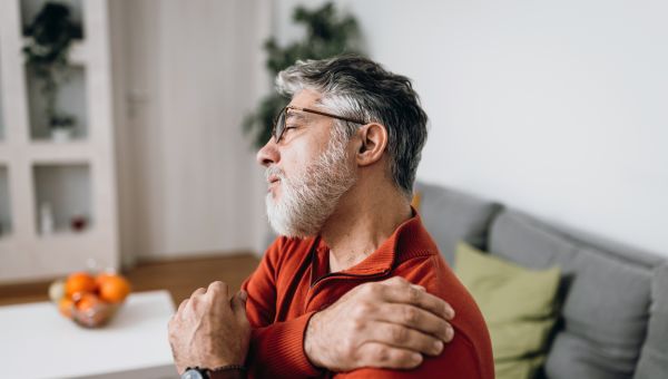 a middle aged white man with a beard sits on his bed and rubs his aching shoulder
