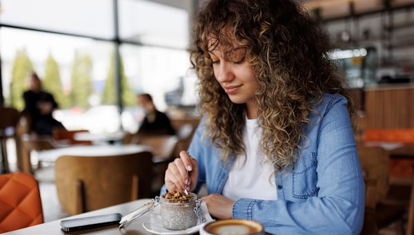 young, curly-haired woman eating chia seed pudding and drinking coffee at a cafe