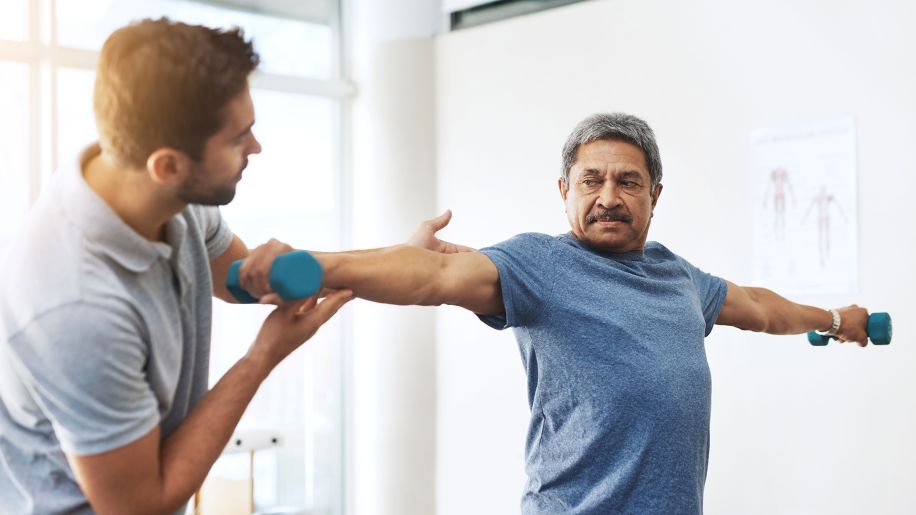 Older man working with a trainer and lifting hand weights while stretching.